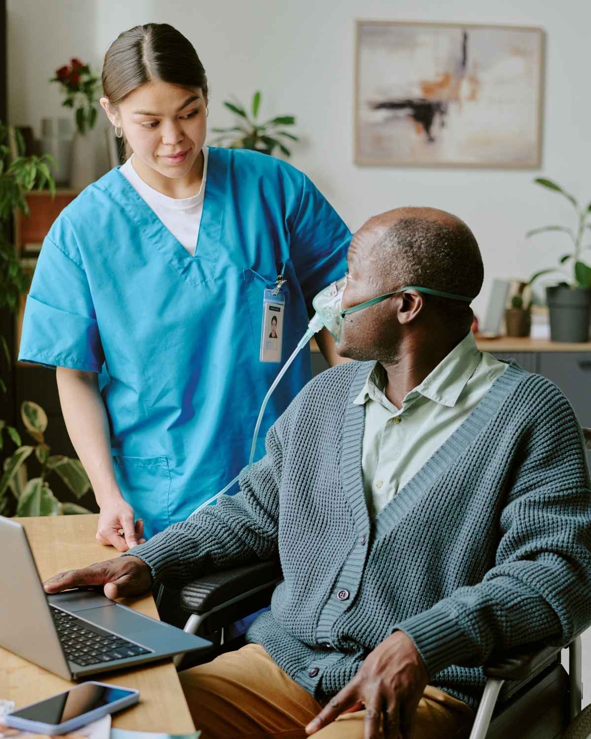 Nurse in blue scrubs assisting a middle-aged man in a wheelchair wearing an oxygen mask while using a laptop.