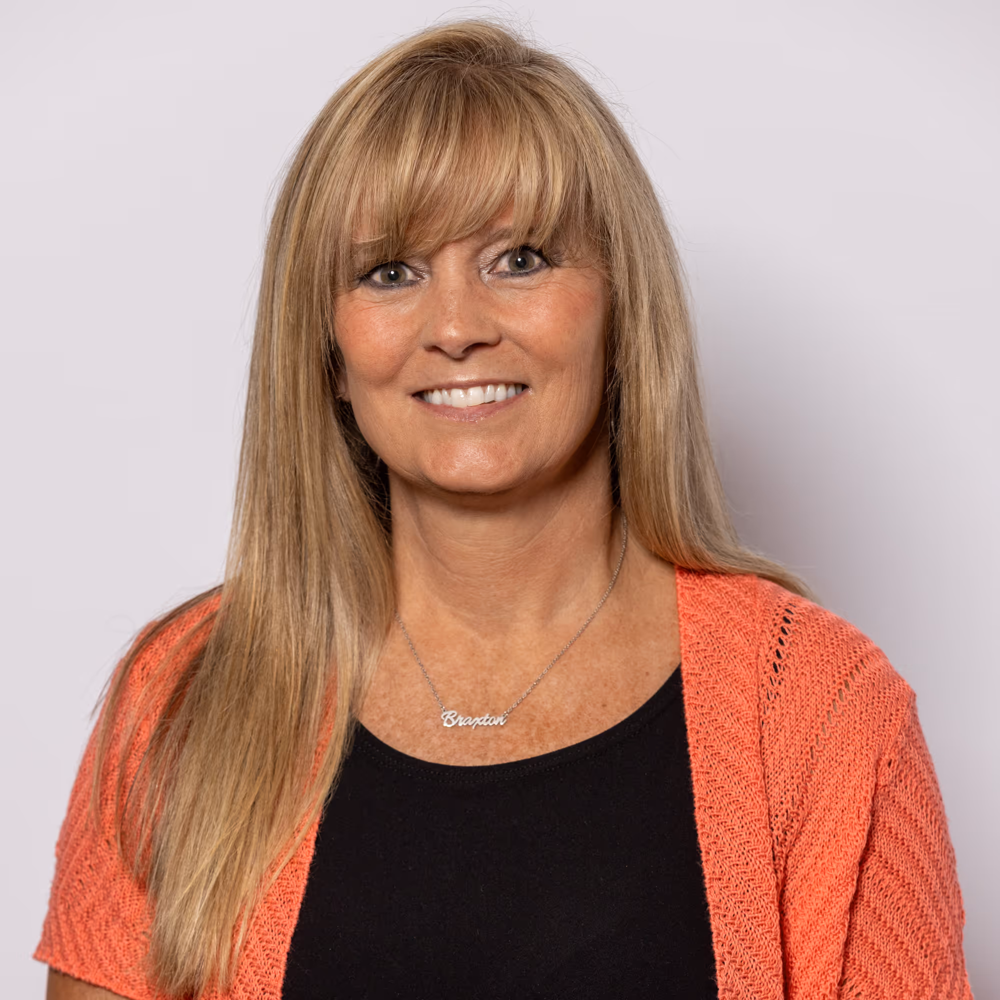 Smiling woman with long blonde hair wearing a black top and coral knit cardigan against a plain light background.
