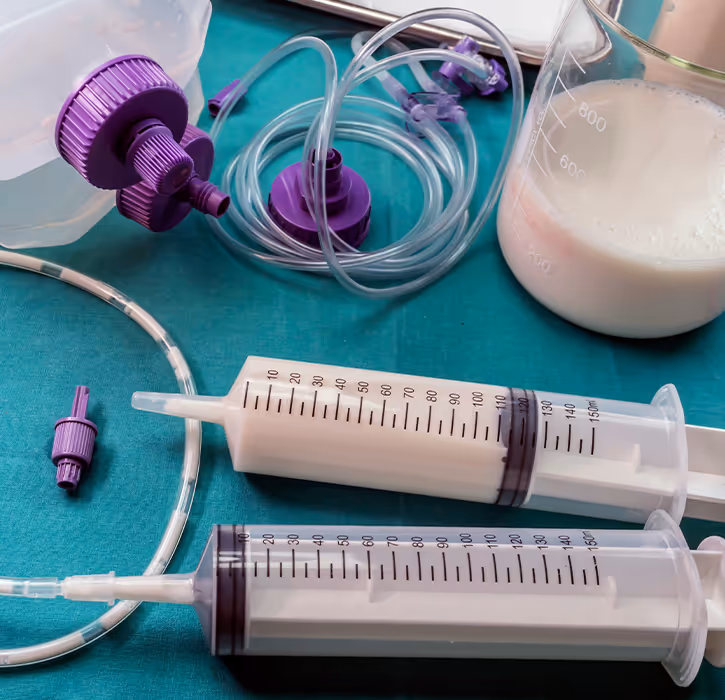 Two large syringes filled with liquid nutrition beside tubing and purple connectors on a teal surface, with a beaker partially filled with milky liquid.