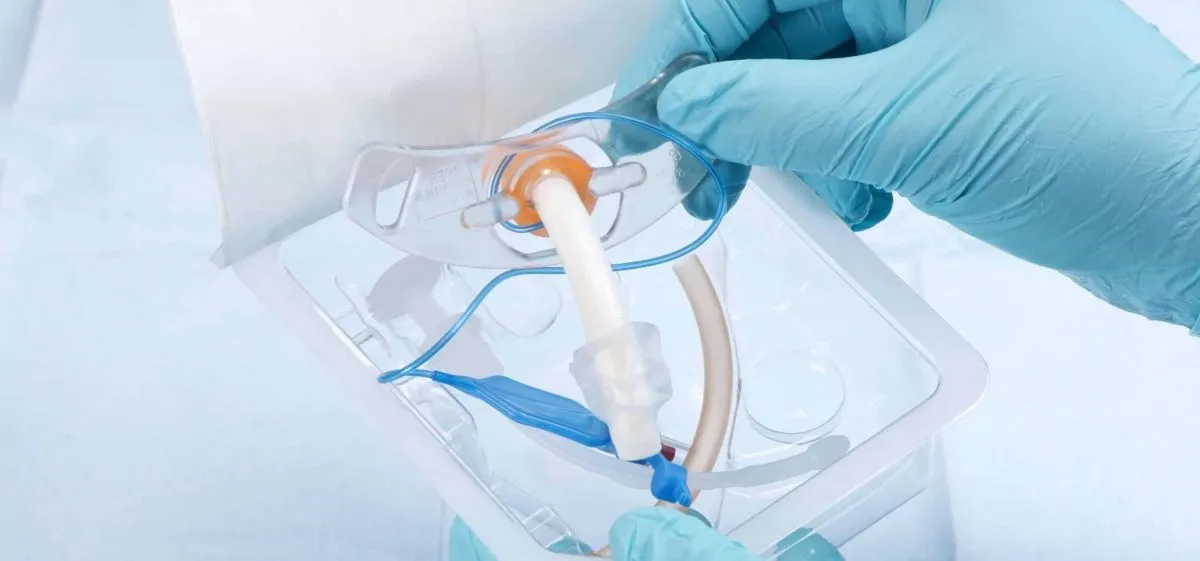 Gloved hands holding a breathing tube with an orange and blue connector inside clear plastic medical packaging.