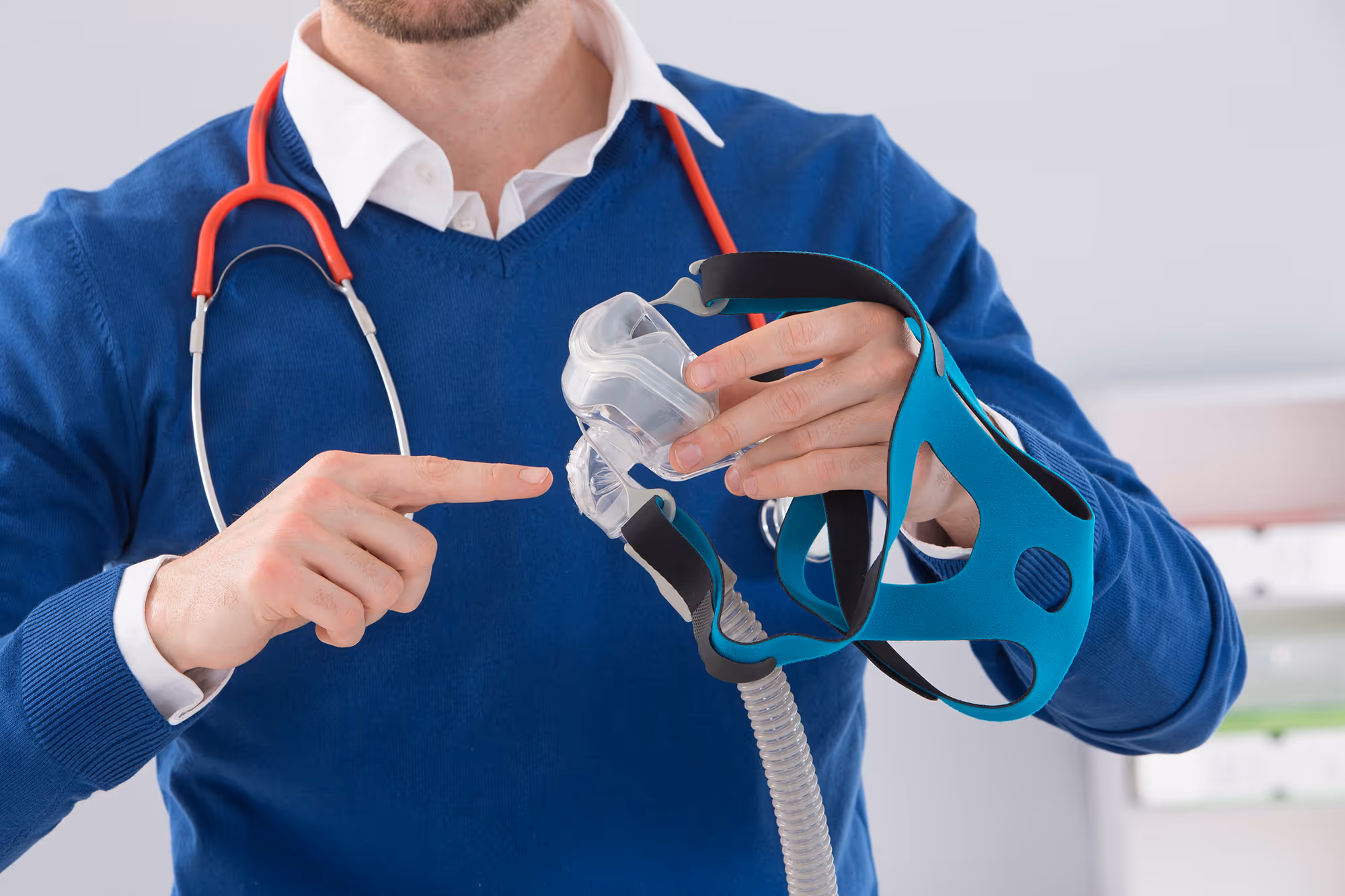 Man in blue sweater with red stethoscope demonstrating a CPAP mask.