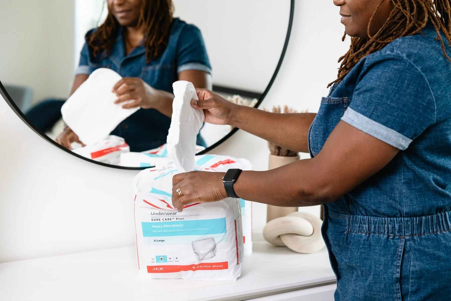 Person holding a white wipe above a package of Sure Care Plus heavy absorbency underwear on a white countertop with a round mirror in the background.