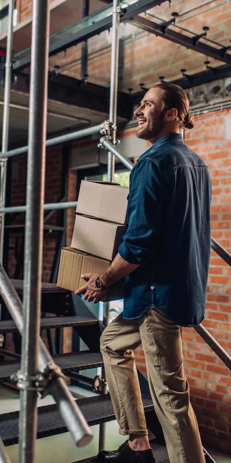 Smiling worker carrying a stack of cardboard boxes up a staircase