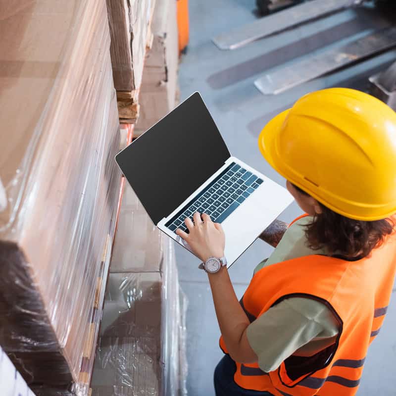 Warehouse worker in a hard hat and hi-vis vest checking inventory on a laptop
