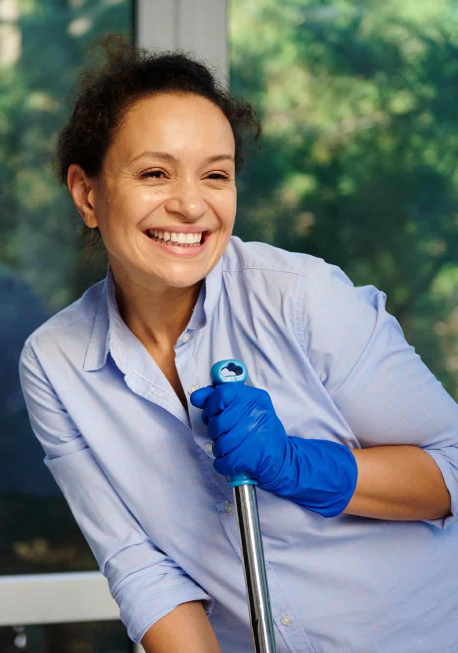 Smiling cleaning worker in gloves leaning on a mop handle by a window