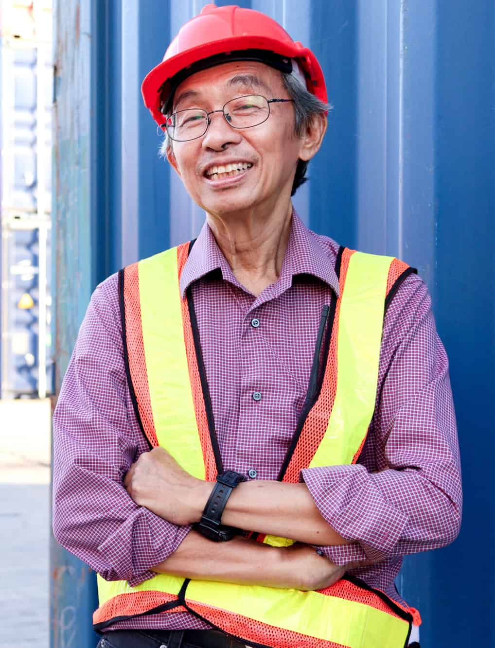 Smiling senior worker in a hard hat and hi-vis vest with arms crossed at a shipping yard