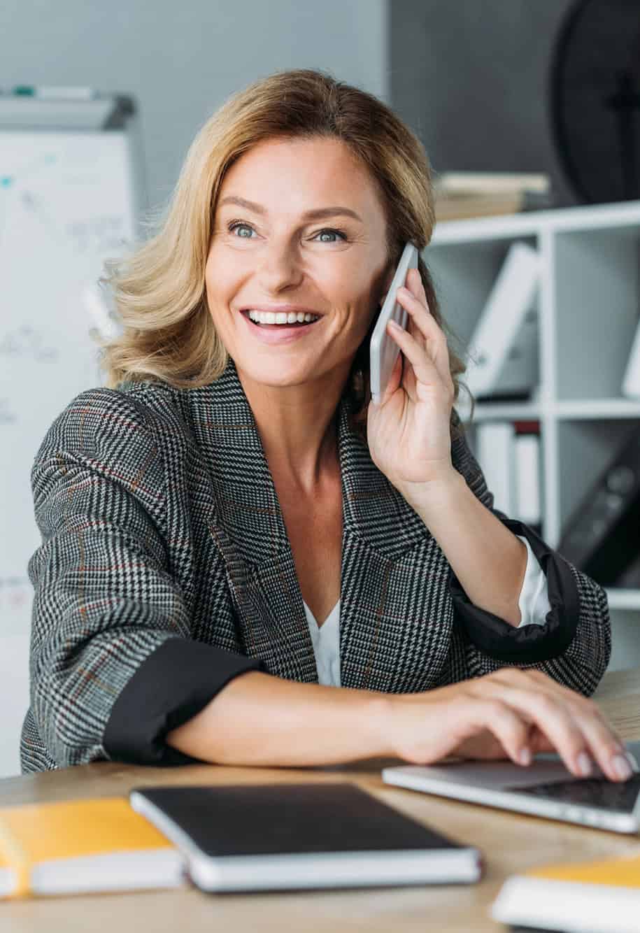 Smiling businesswoman in a blazer talking on a smartphone at her office desk
