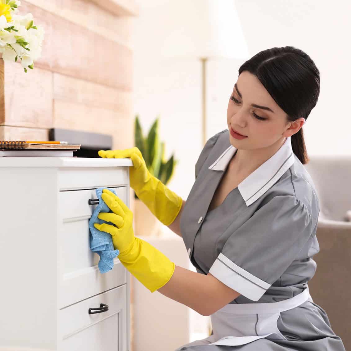 Housekeeper in uniform and gloves dusting furniture in a hotel room