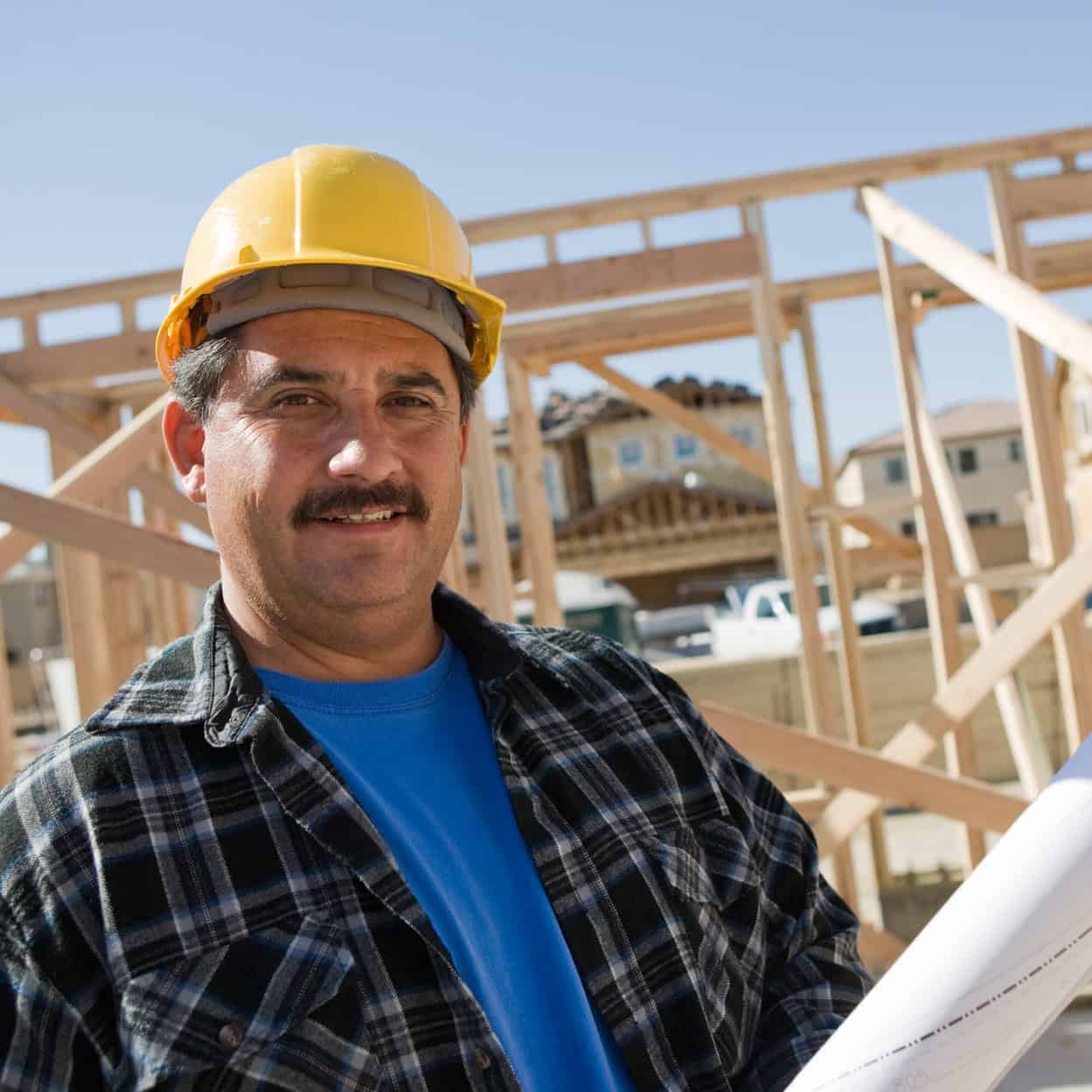 Smiling construction worker in a hard hat holding blueprints at a house framing site