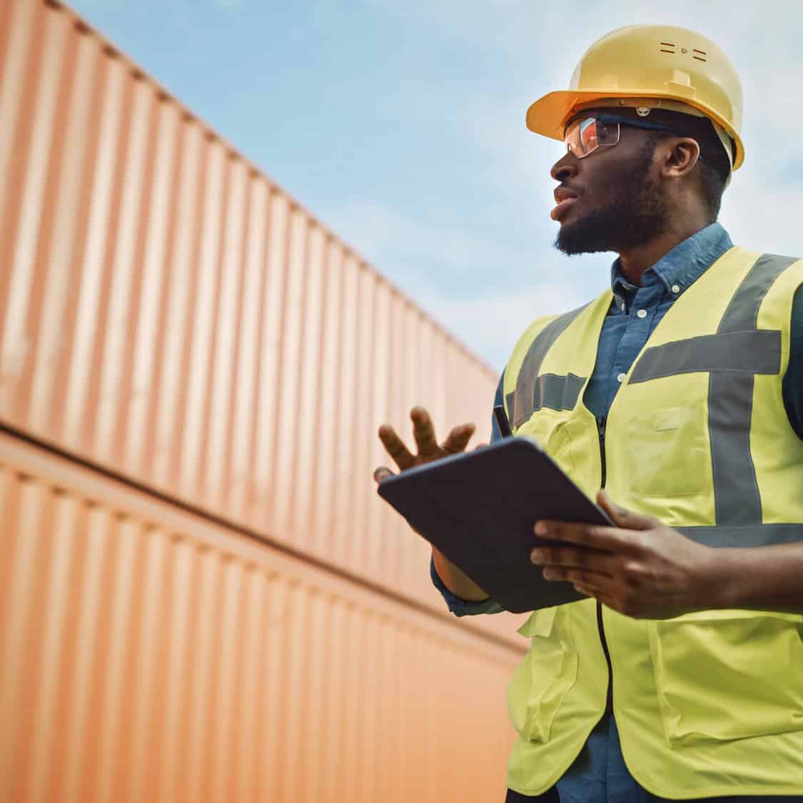 Logistics worker in a hard hat and hi-vis vest holding a tablet beside shipping containers