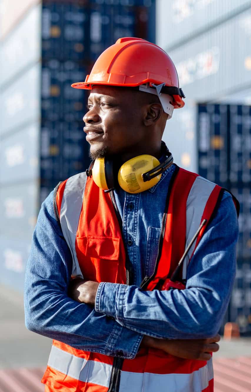 Confident logistics worker in a hard hat and safety vest with arms crossed at a shipping container yard