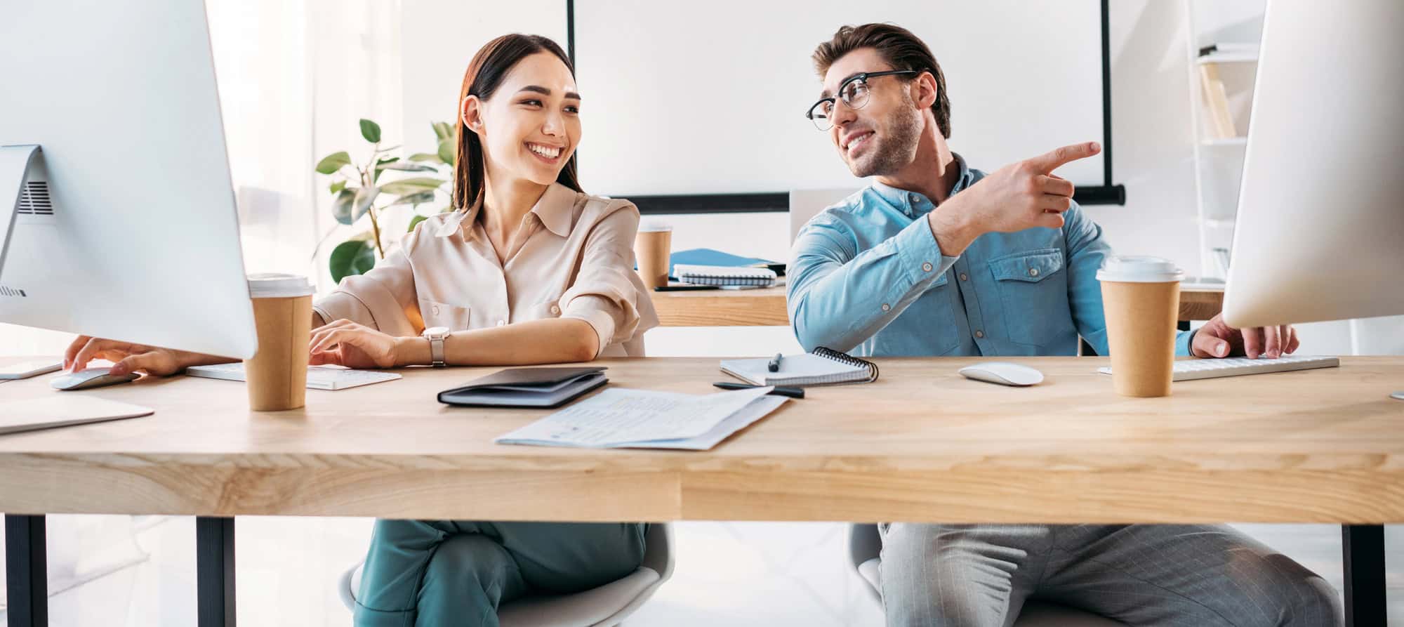 Two smiling coworkers collaborating at desktop computers in a bright office
