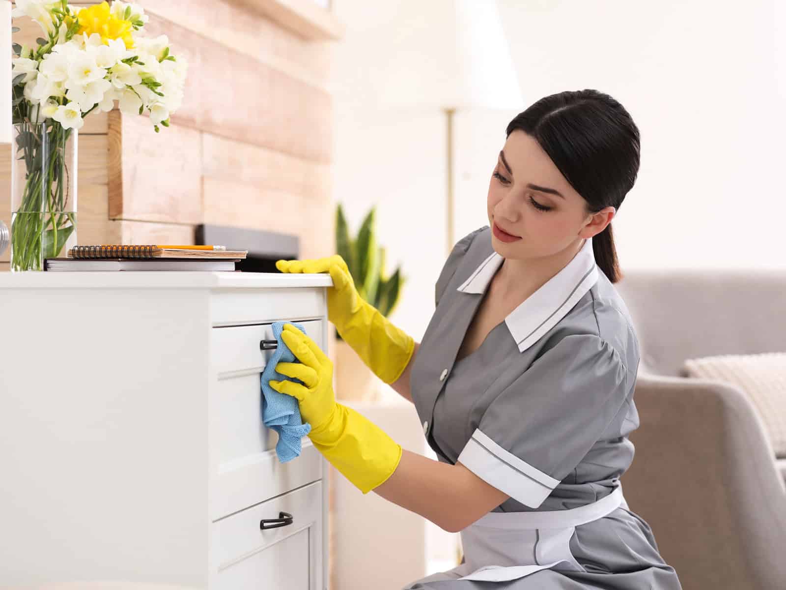 Housekeeper in uniform and gloves dusting furniture in a hotel room