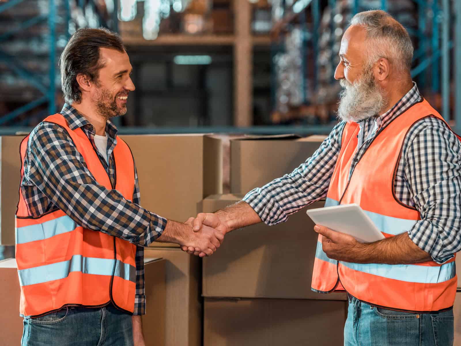Two warehouse workers in safety vests shaking hands among stacked boxes