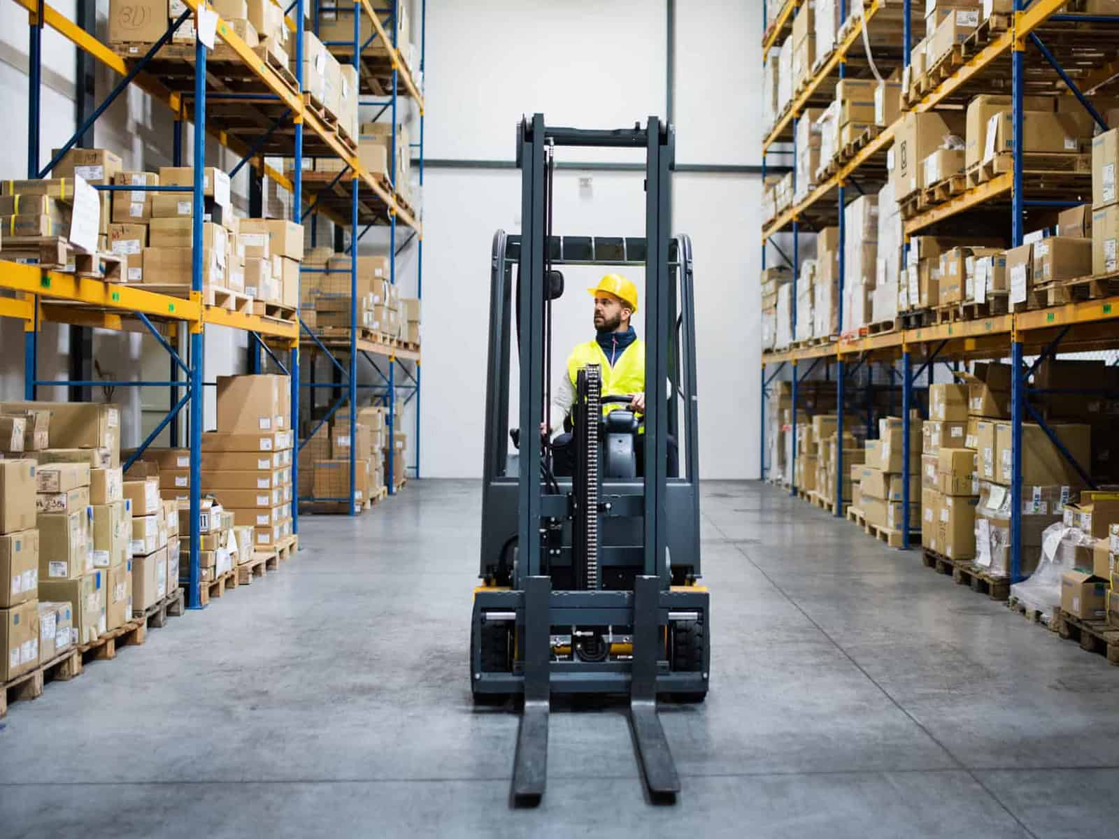 Forklift operator in a hard hat and hi-vis jacket driving down a warehouse aisle