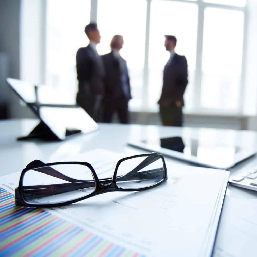 Glasses and reports on a desk with business colleagues talking in the background