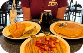 Table with a breakfast burger, two plates of pancakes with syrup and strawberries, and a person wearing a maroon Deejay’s diner shirt in the background.