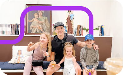 Five children smiling while sitting on a couch with a large framed painting and bookshelf in the background.