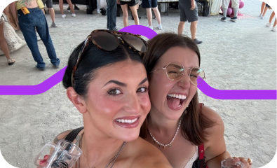 Two women smiling and holding drinks at an outdoor event with other people in the background.