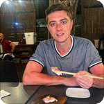 Young man smiling at a restaurant table holding chopsticks with sushi in front of him.