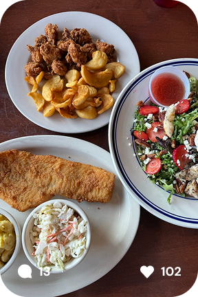 Two plates of food on a wooden table, one with fried chicken pieces and potato wedges, and another with a fresh salad and dipping sauce, accompanied by a third plate with fried fish and coleslaw.