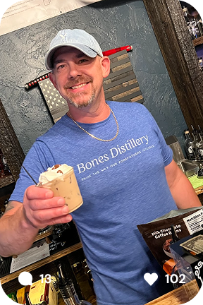 Man wearing a Bones Distillery t-shirt and cap smiling behind a bar, holding a whipped cream topped iced coffee.