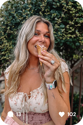 Smiling woman with long blonde hair enjoying an ice cream cone outdoors in front of greenery.