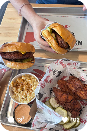 Tray with two fried chicken sandwiches, a serving of macaroni and cheese, pickles, and dipping sauce, one sandwich held by a hand.