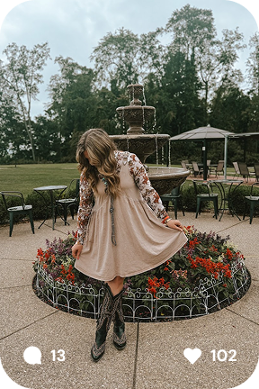 Woman wearing a beige dress and cowboy boots posing in front of a flower bed and fountain outdoors.