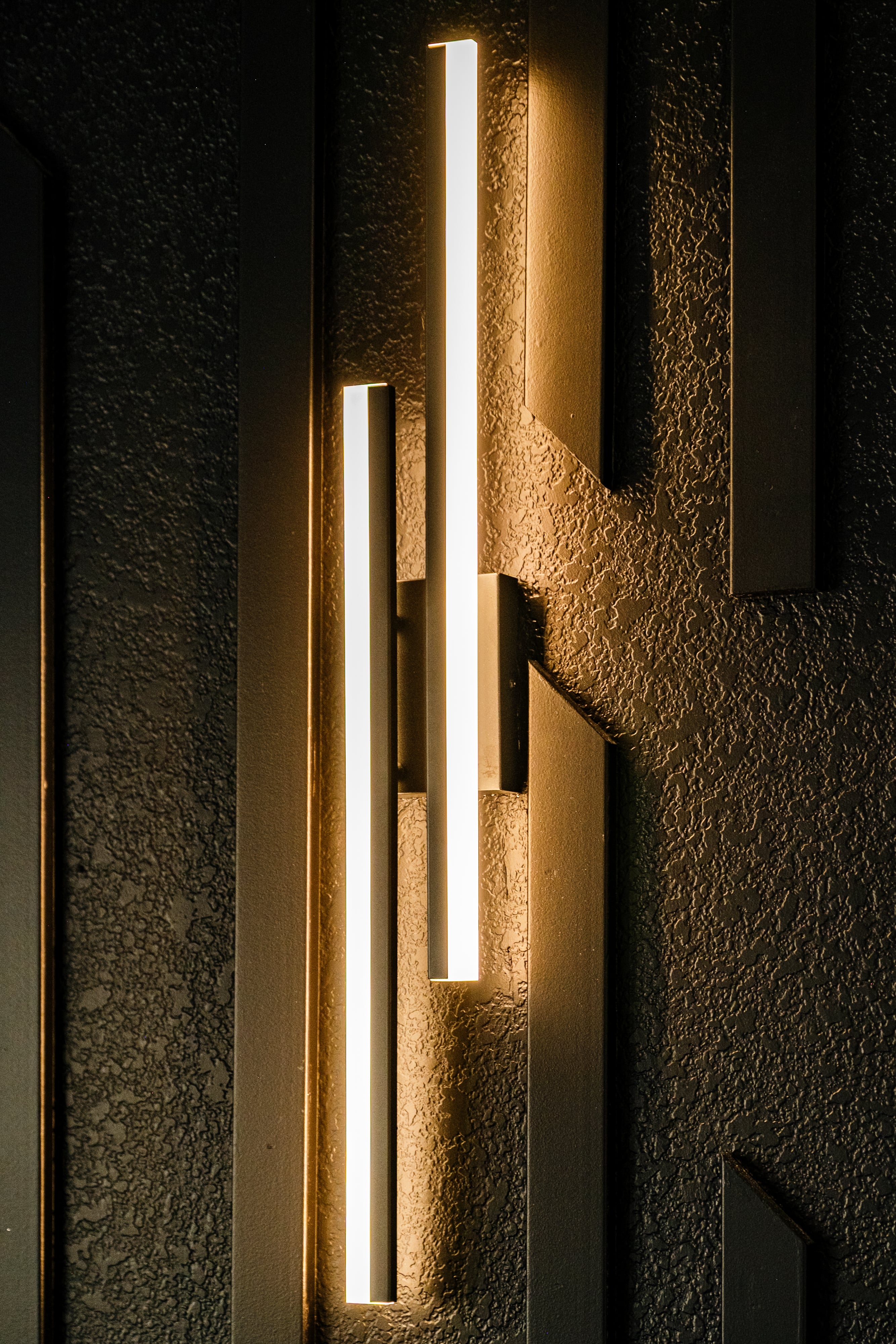 Modern wall light fixture with two vertical glowing bars mounted on textured dark wall panels.