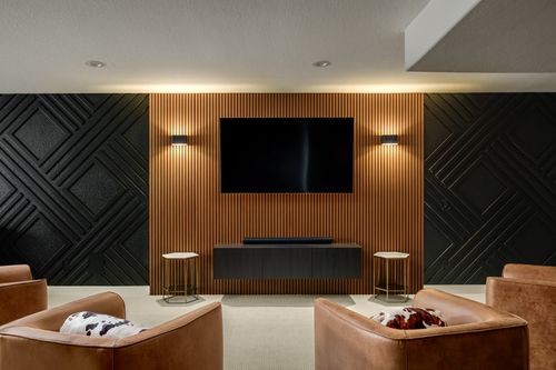 Modern living room with tan leather chairs, a large flat-screen TV mounted on a vertical wooden slat wall, black textured accent walls, and small marble-topped side tables.