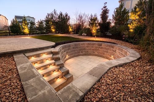 Stone sunken patio with built-in stairs illuminated by lights surrounded by landscaping with small trees and rocks at dusk.