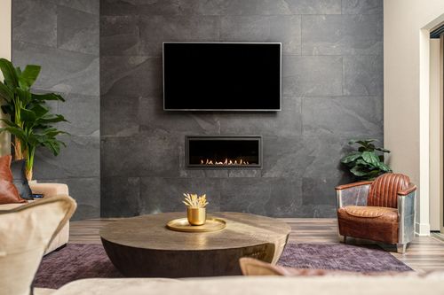 Modern living room with dark stone wall, wall-mounted TV, built-in fireplace, round wooden coffee table, leather chair, and indoor plants.