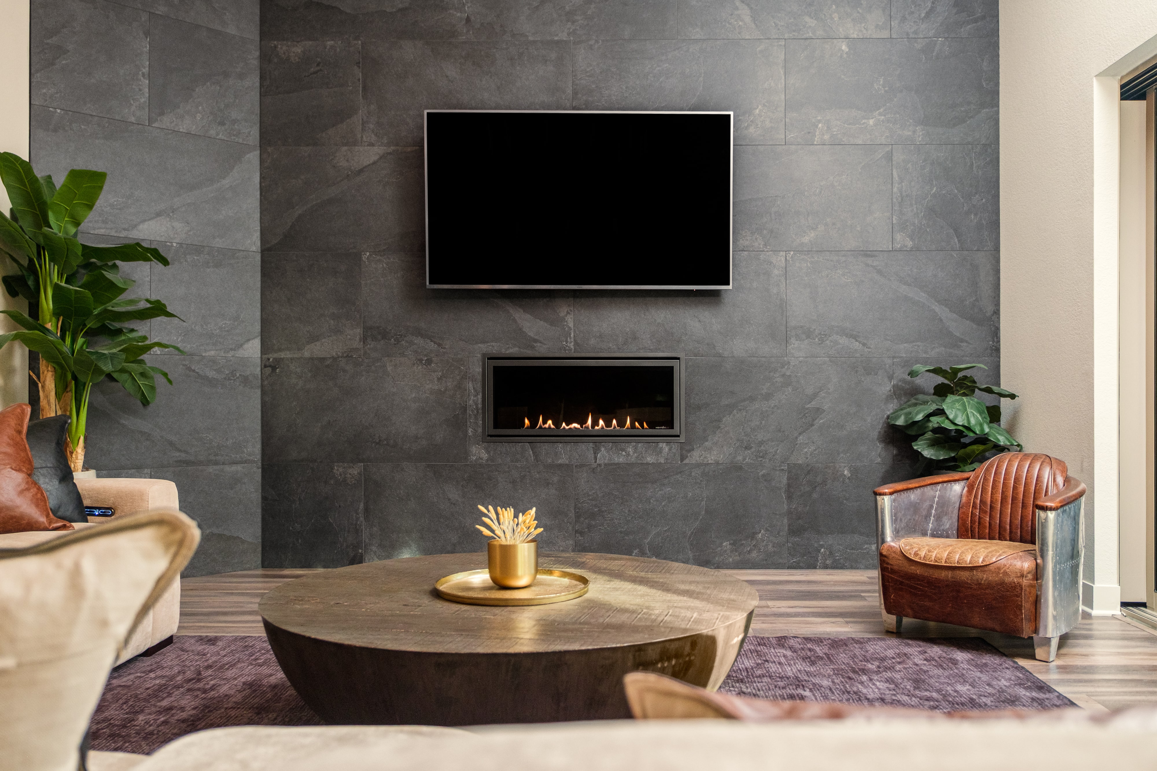 Modern living room with dark stone wall, wall-mounted TV, built-in fireplace, round wooden coffee table, leather chair, and indoor plants.