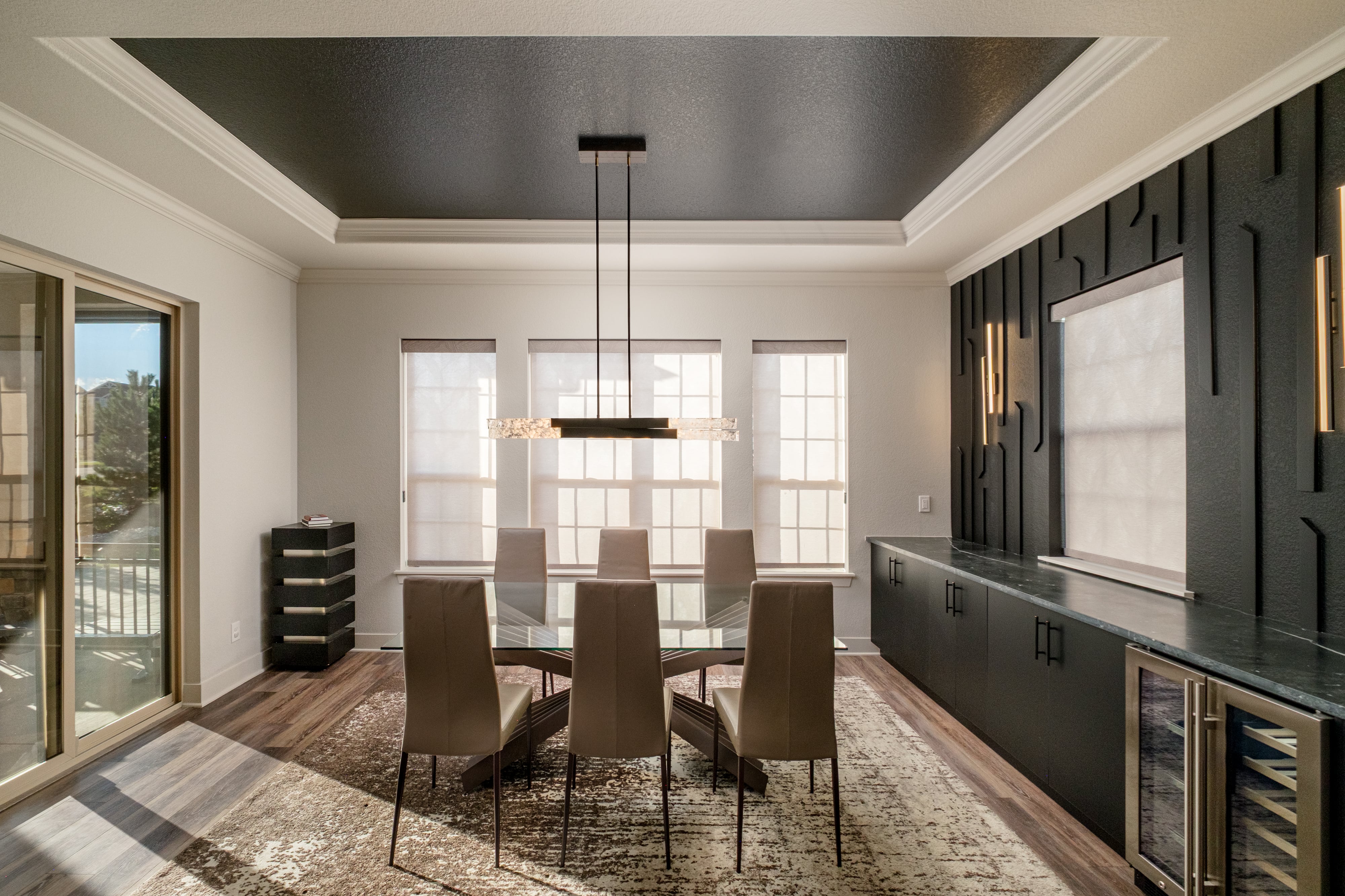 Modern dining room with a glass table, six beige chairs, black accent wall with built-in cabinets, and large windows with shades.