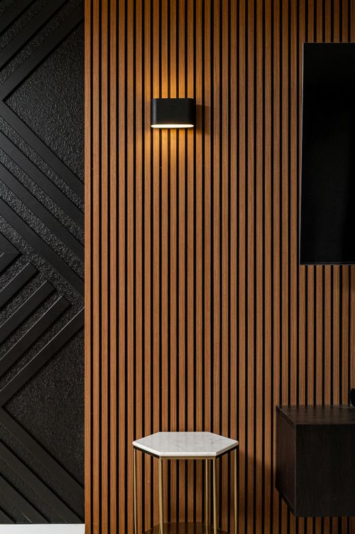 Modern interior wall with vertical wooden slats, illuminated black wall sconce, hexagonal marble-top table, and part of a black cabinet and TV screen.