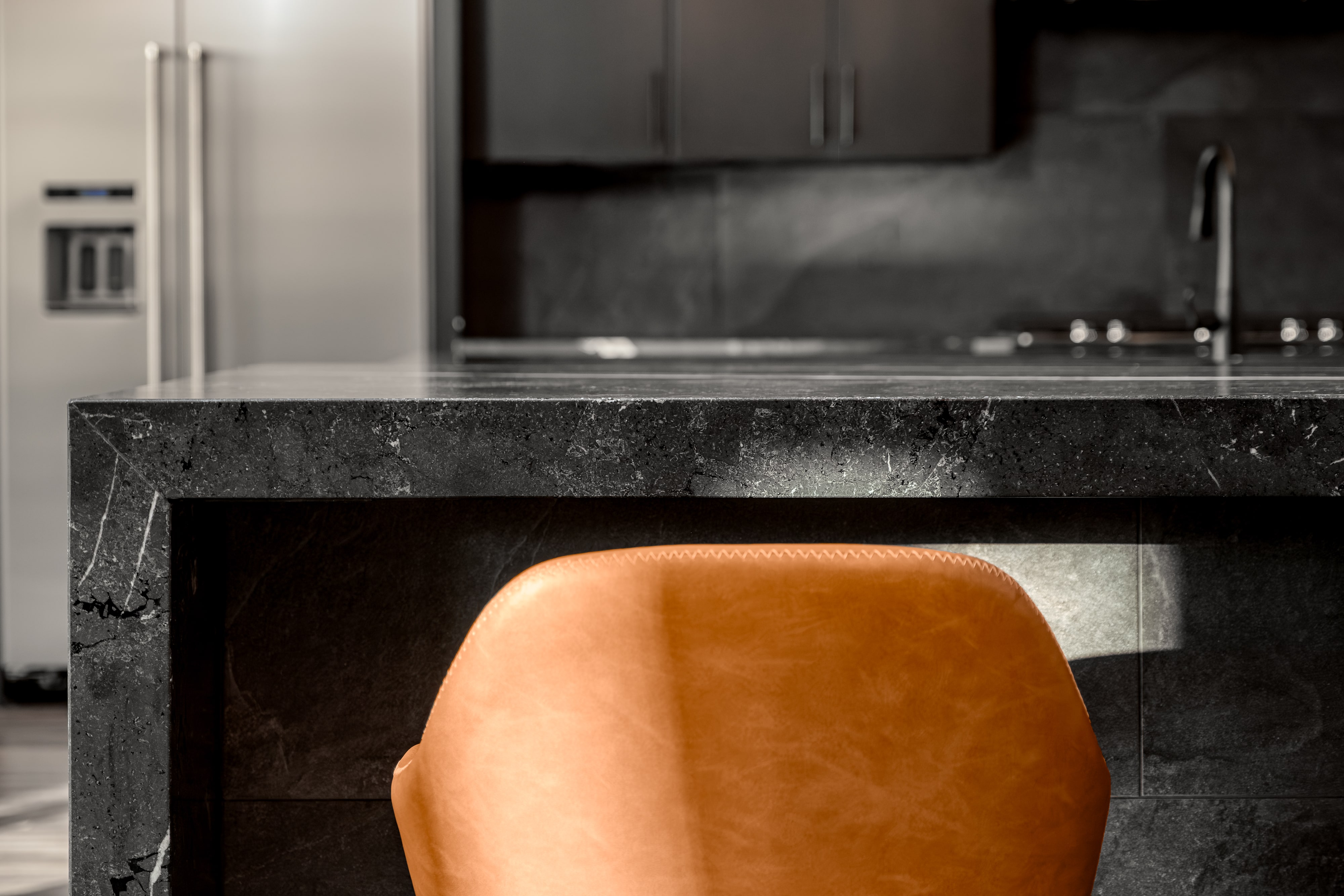 Close-up of a tan leather chair back with a black stone kitchen island and stainless steel appliances in the background.