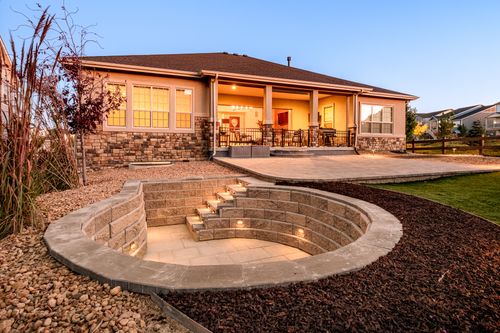 Backyard of a house at dusk with stone exterior walls, a covered patio, circular sunken seating area with built-in lights, and landscaped garden beds.