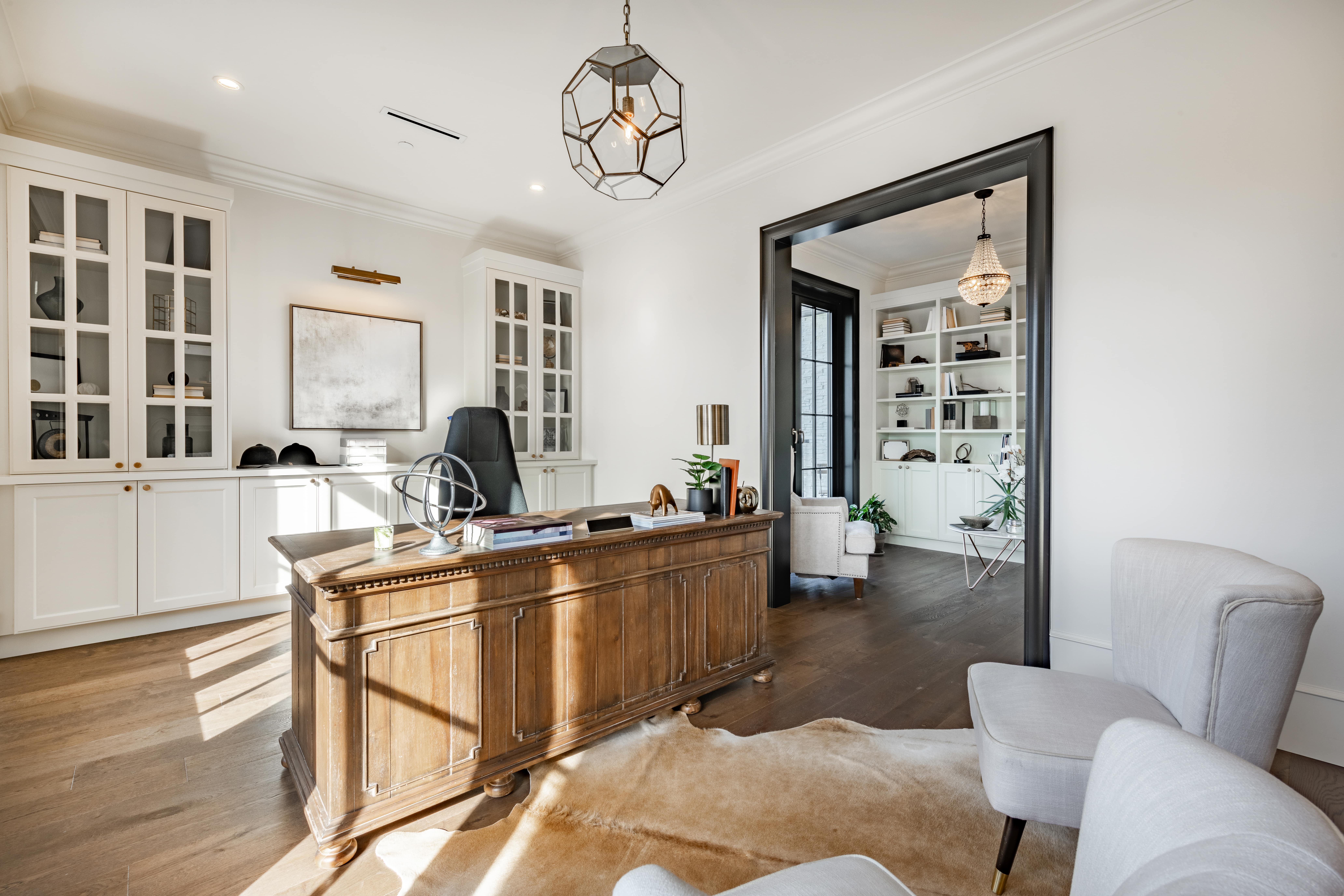 Bright home office with a wooden desk, black chair, white armchairs, and built-in shelves with decorative items.