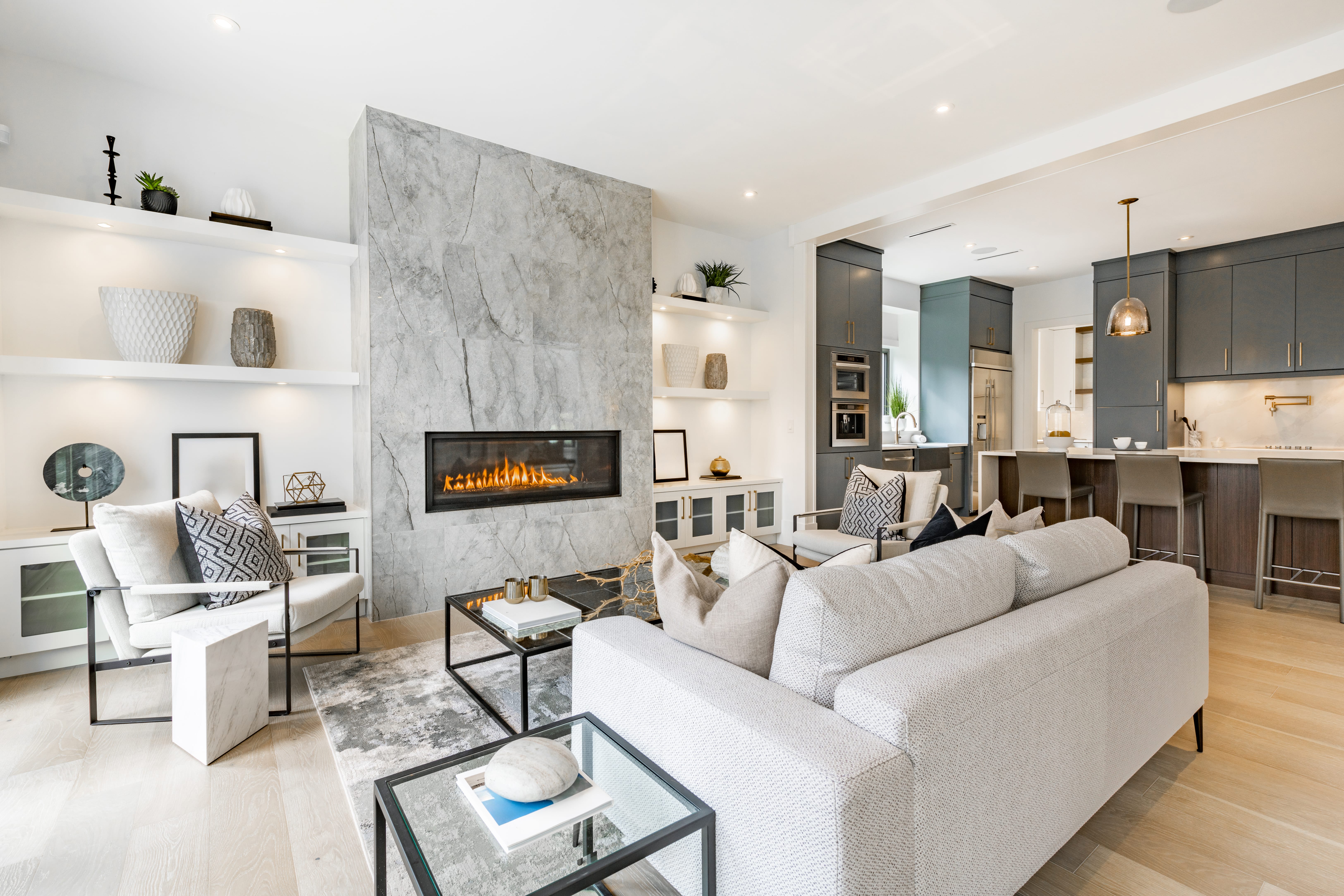 Bright modern living room with light gray sofa, armchairs, a rectangular gas fireplace set in a marble wall, and an open kitchen with dark cabinets and bar stools.
