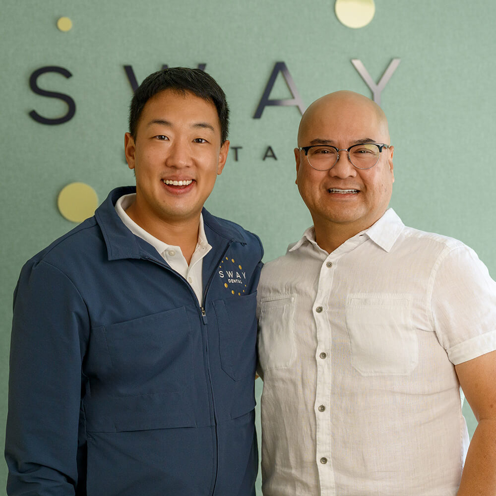 Two men smiling in front of a green wall with a partial SWAY DENTAL logo, one wearing glasses and a white shirt and the other in a blue jacket with the same logo.