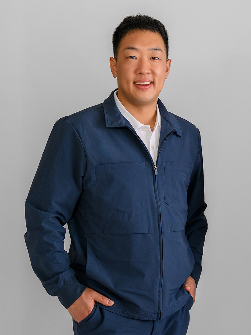 Smiling man wearing a navy blue jacket and white shirt standing against a light gray background with hands in pockets.