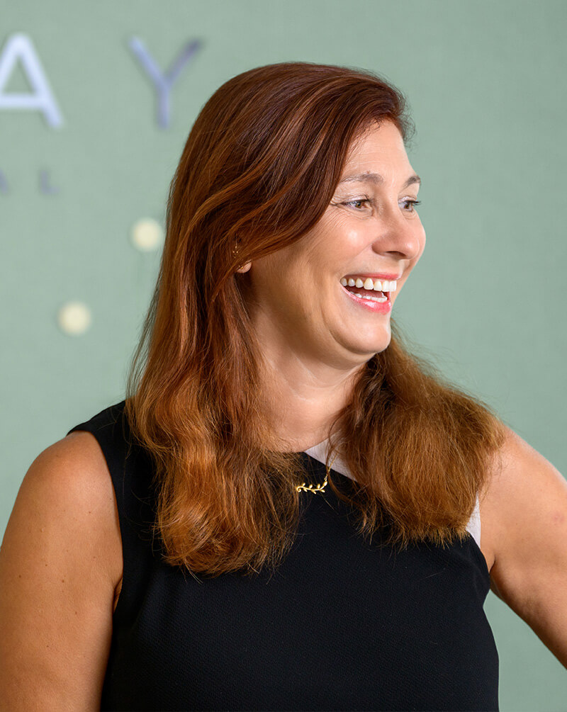 Smiling woman with long reddish hair wearing a black sleeveless top and a gold necklace, looking to the right.