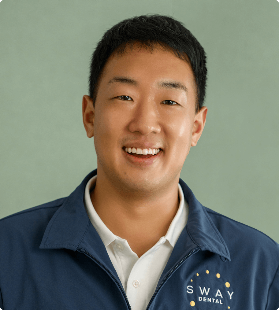 Smiling man wearing a navy jacket with SWAY Dental logo over a white polo shirt, with a muted green background.