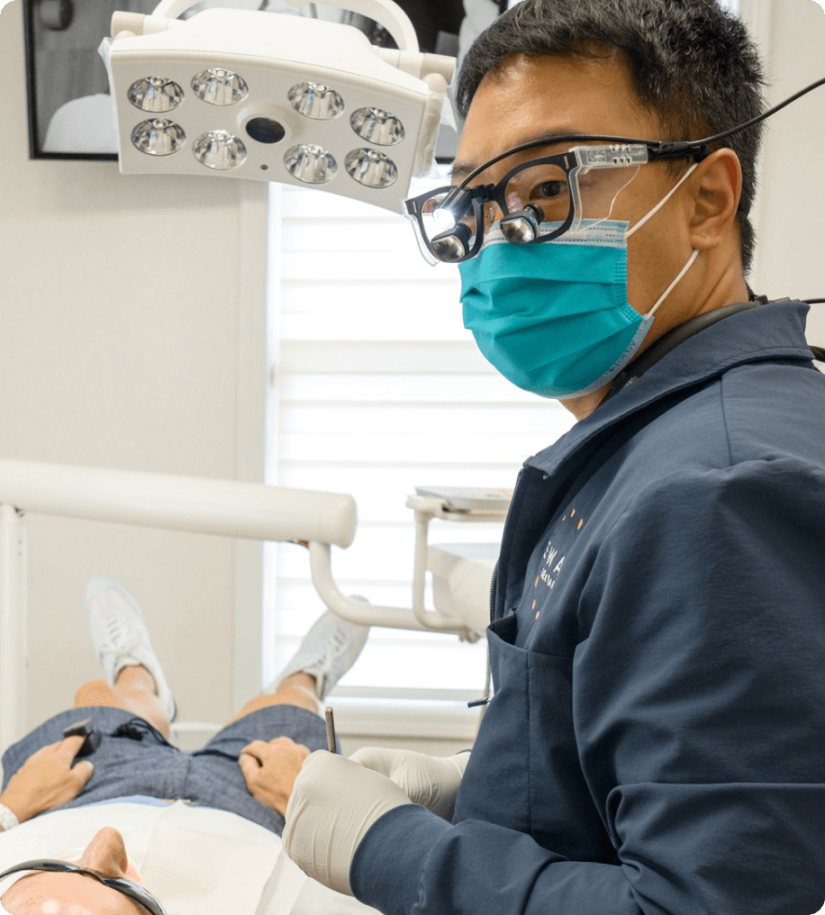 Dentist wearing magnifying glasses and a blue surgical mask examining a patient lying in a dental chair.