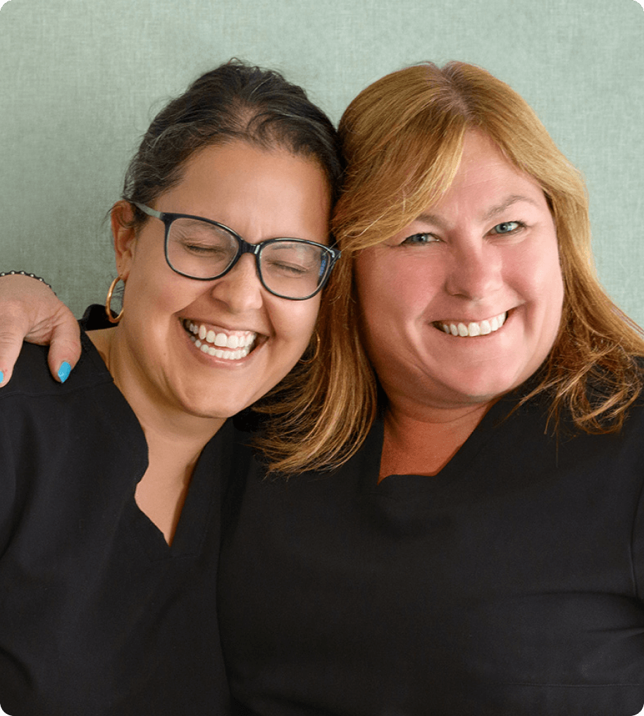 Two women smiling warmly, one with glasses and closed eyes, the other with blonde hair; both wearing black tops.
