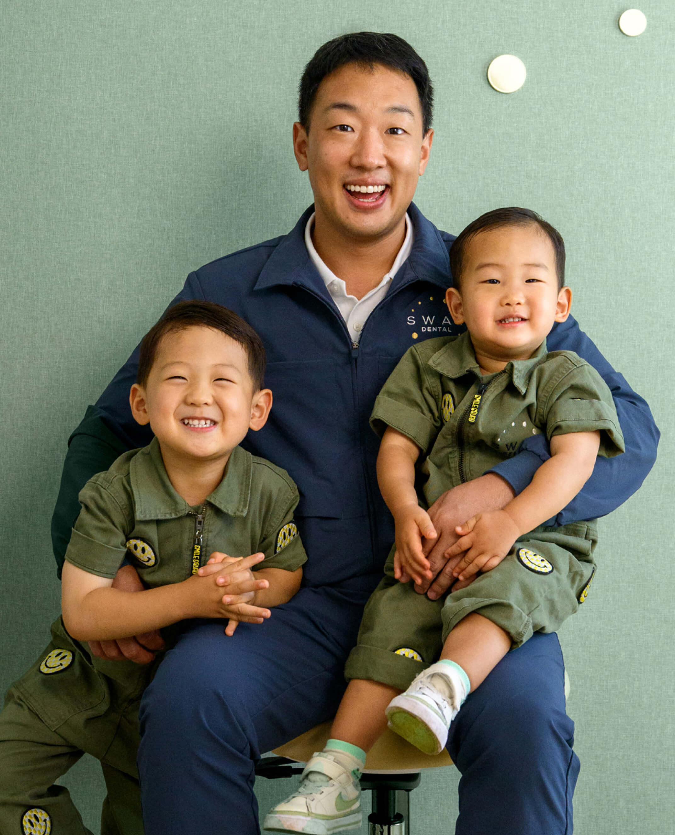 Smiling man in navy jacket with two young boys wearing matching green outfits with smiley face patches, posed against light green wall.