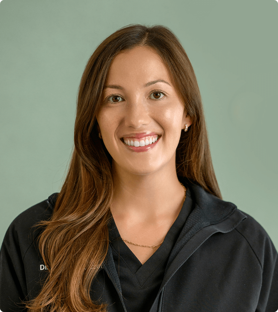 Smiling woman with long brown hair wearing a black jacket over a black top against a muted green background.