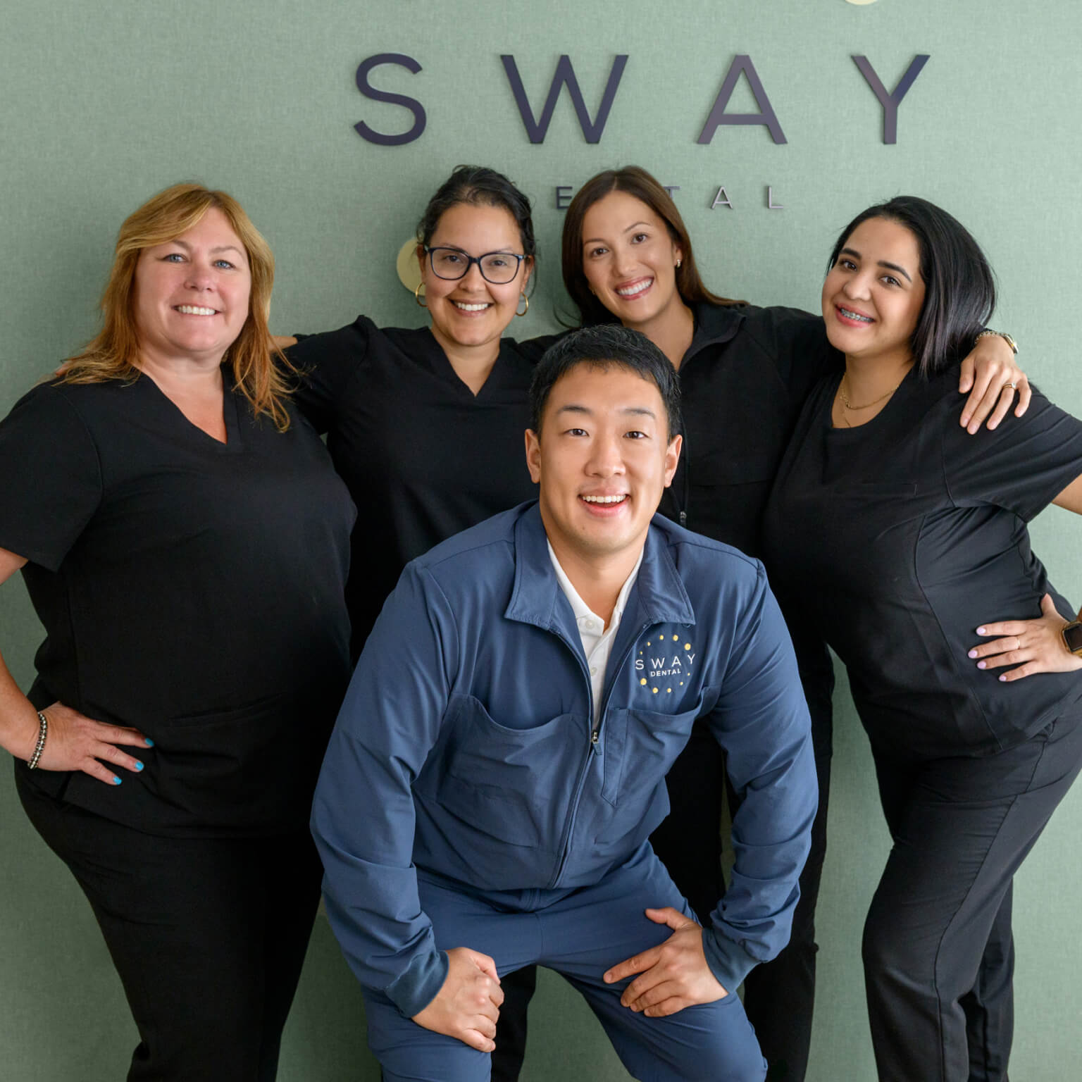 Five dental professionals smiling and posing in front of a wall with the SWAY Dental logo, four in black uniforms and one in a blue SWAY Dental jacket.