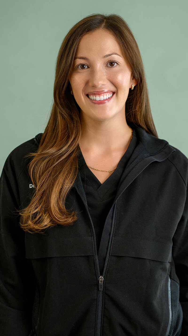 Smiling woman with long brown hair wearing a black zip-up jacket and a delicate gold necklace against a light green background.